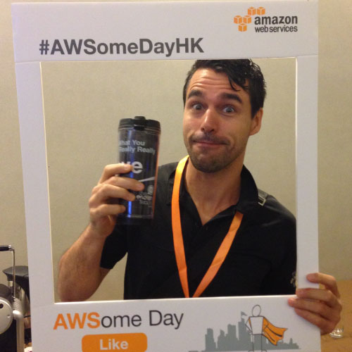 A man lifting his coffee tumbler towards the photographer as in saying: Cheers! whilst posing in front of a foamboard frame that says AWSomeday by Amazon.