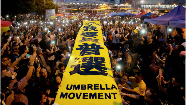 photo of thousands of Hongkongers at night lighting their mobile phone flashes and stretching a yellow banner over the heads of hunderes of people and with bold black words both in English and traditional Chinese: Umbrella Movement