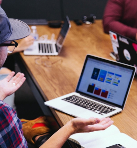 a man with a cap and glasses explaining or questioning ideas in a brainstorm on a wooden table with a few laptops open